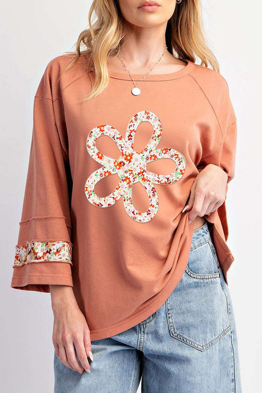 Flower Patch Graphic Top in orange with floral applique flower and exposed seams, styled with light denim jeans for casual spring wear.
