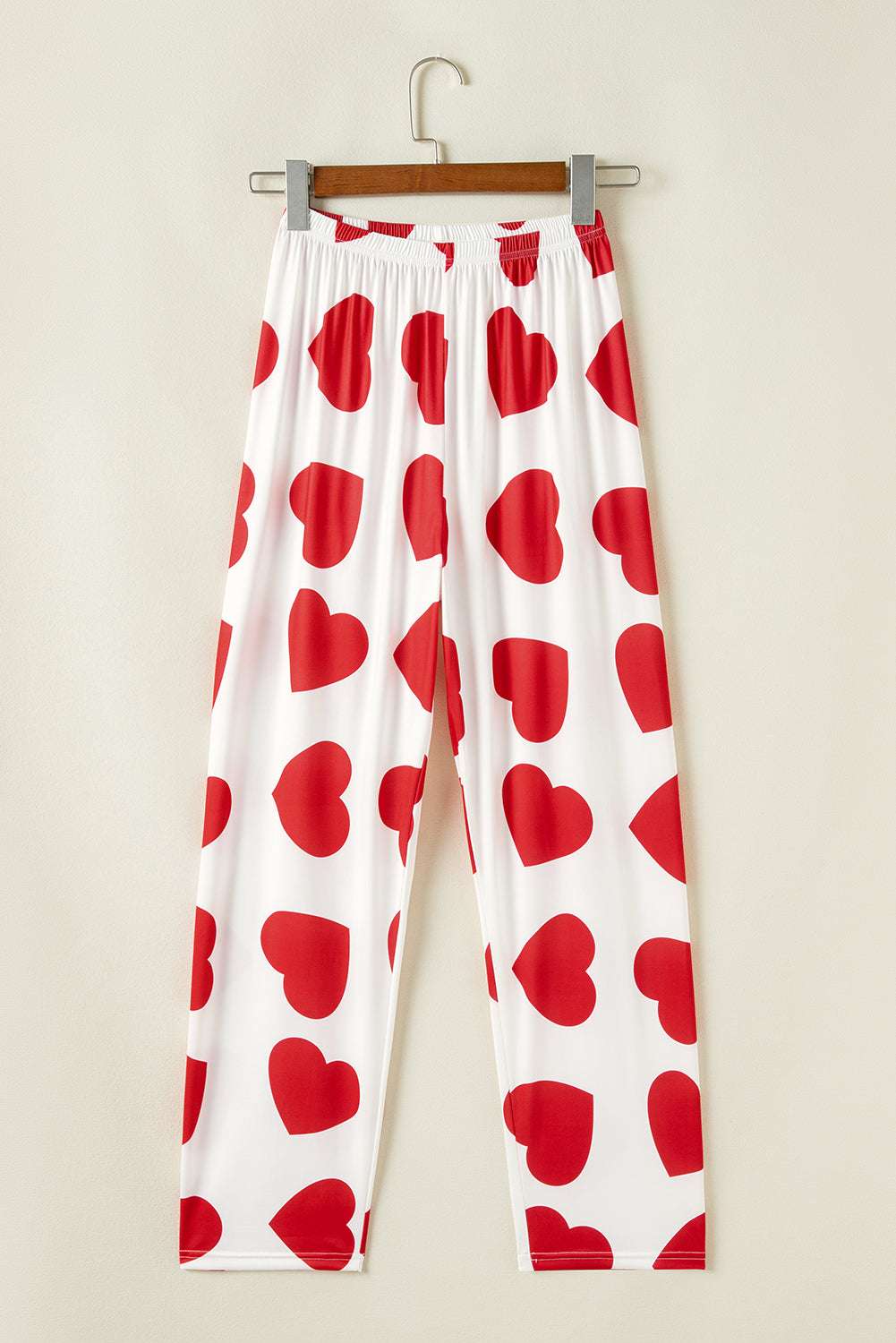 White pants with red heart patterns hanging on a wooden hanger against a beige wall.