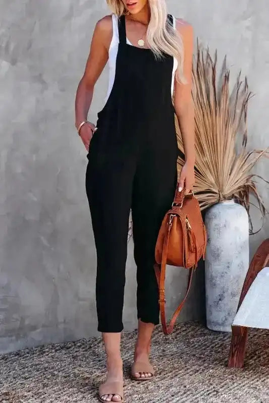 Black cropped jumpsuit with adjustable buckle straps styled with a white tank top and tan handbag. Sleeveless and ideal for casual wear.