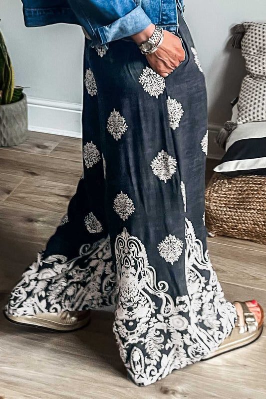 Women's black geometric print wide leg trousers in plus size with paisley border, styled casually with sandals and denim jacket indoors.