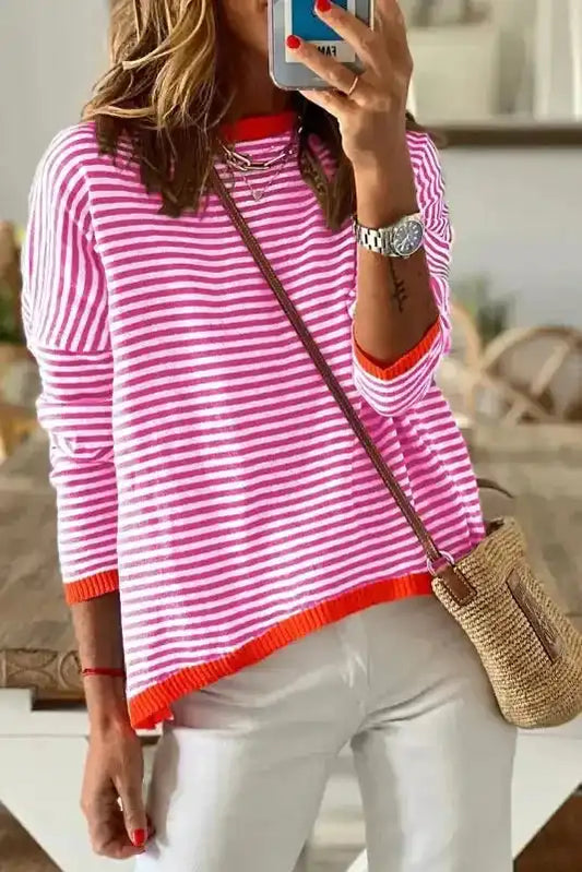 Women's striped baggy sweater in pink with red contrast trim, styled casually with white pants and a straw crossbody bag indoors.