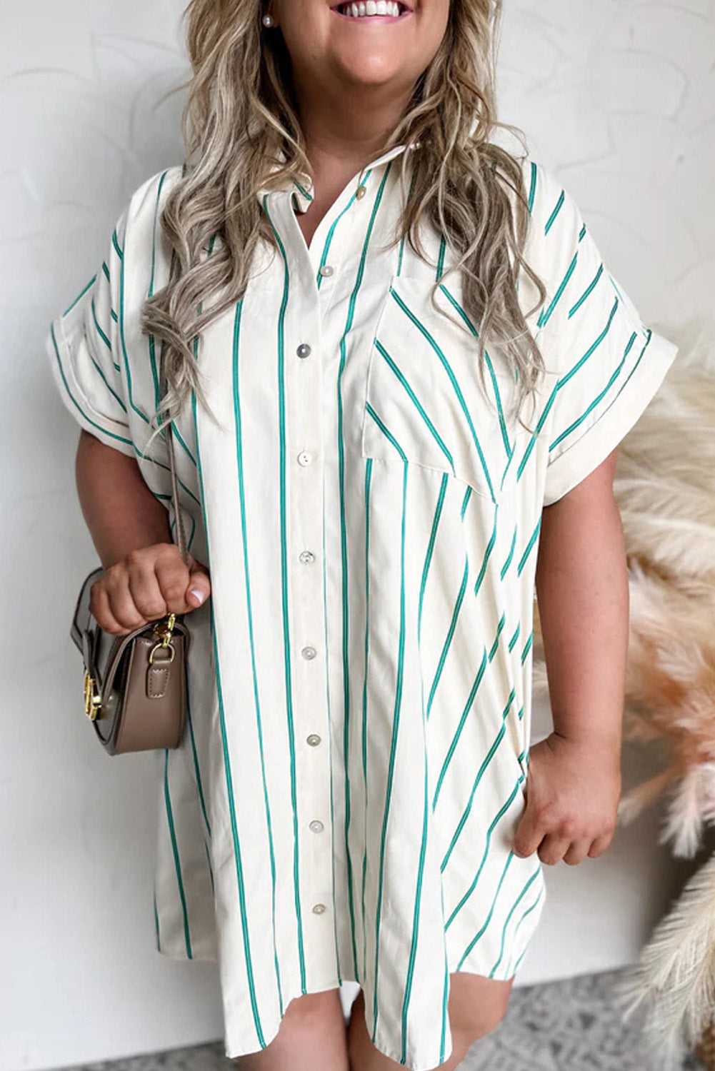 Woman wearing a white shirt with green stripes, holding a beige handbag against a neutral background.