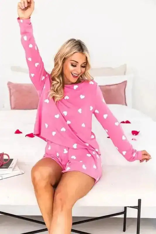 Woman wearing Pink Valentine Lounge Set, sitting on bed in heart print tee and shorts, showcasing comfy and festive loungewear.