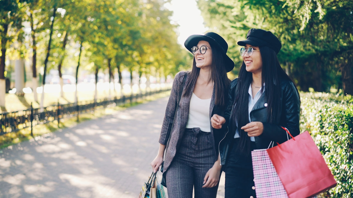 Two women walk with shopping bags in a park.
