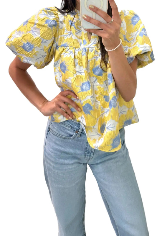 Person wearing a yellow floral blouse and light blue jeans, taking a mirror selfie.