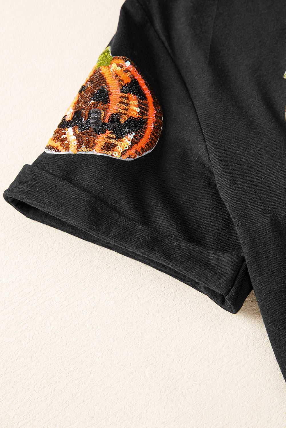 Sequin Pumpkin Face Halloween T-Shirt sleeve close-up with a decorative patch resembling a jack-o'-lantern on a beige background