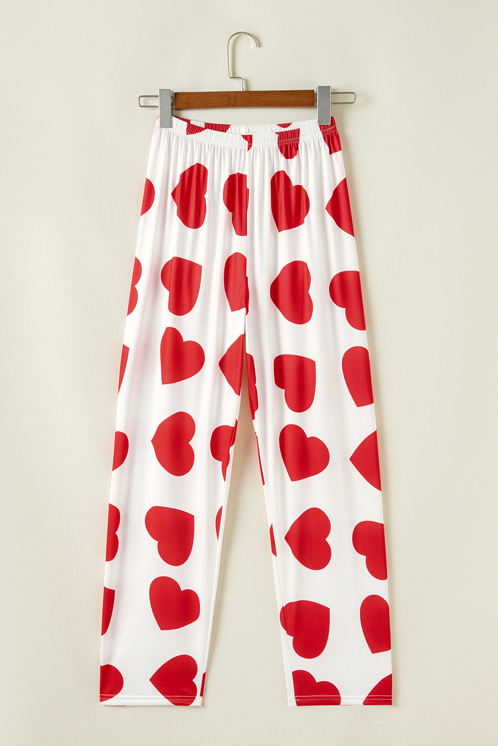White pants with red heart patterns hanging on a wooden hanger against a beige wall.