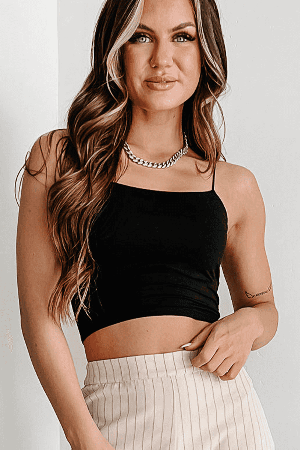 Model wearing black spaghetti strap crop top with stretch fit, styled with neutral pinstripe pants for a modern casual look.