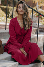Seated model wearing Ladies Burgundy Long Sleeve Tiered Maxi Dress, highlighting soft fabric and elegant layered design.