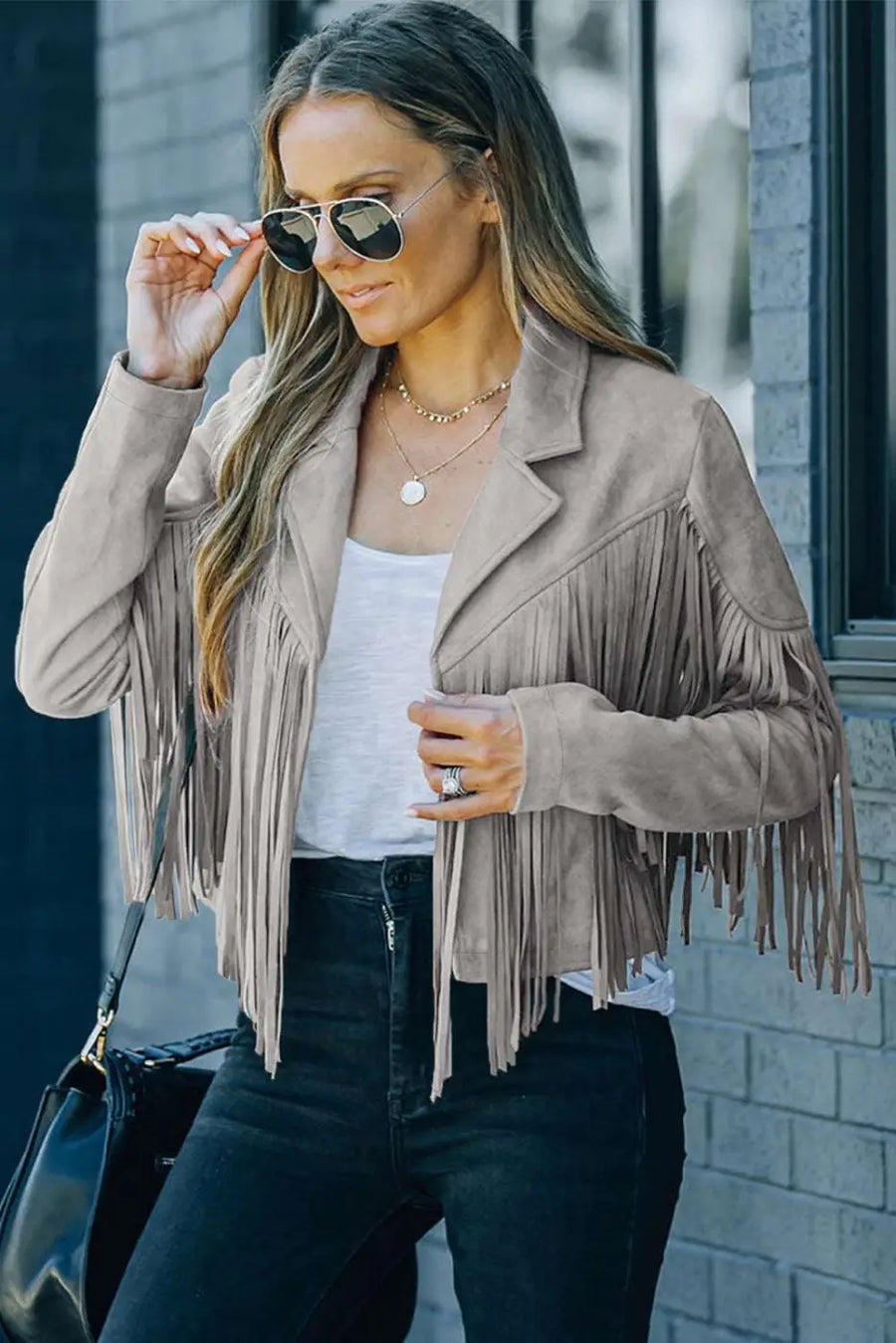 Front view of ladies gray cropped fringe faux suede jacket styled with layered necklaces and black jeans for a chic fall look.