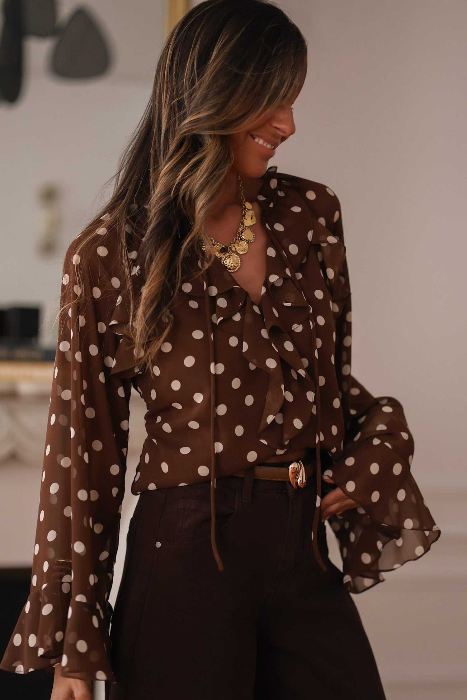 Coffee polka dot bell sleeve blouse for women with tie neck and ruffled details, styled with high-waisted trousers for an elegant office look.