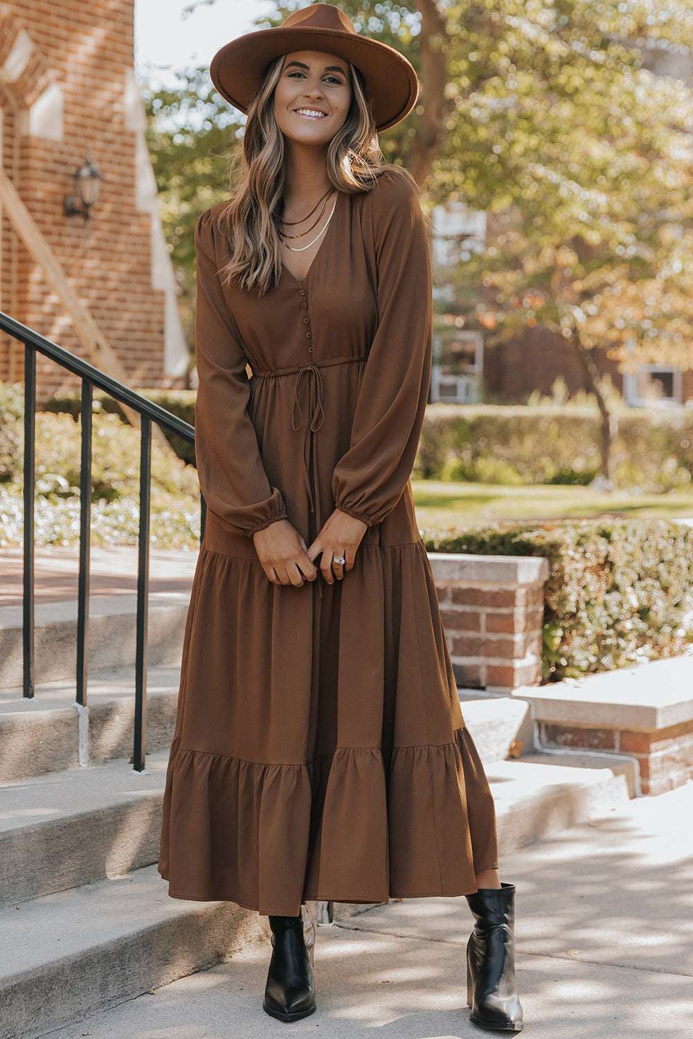 Full front view of Brown Long Sleeve Tiered Maxi Dress with modern A-line cut and cinched waist, styled with a wide-brim hat.