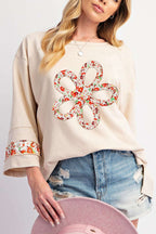 Beige Flower Graphic Top with vibrant floral applique on front and sleeves, styled with distressed denim shorts for casual spring wear.