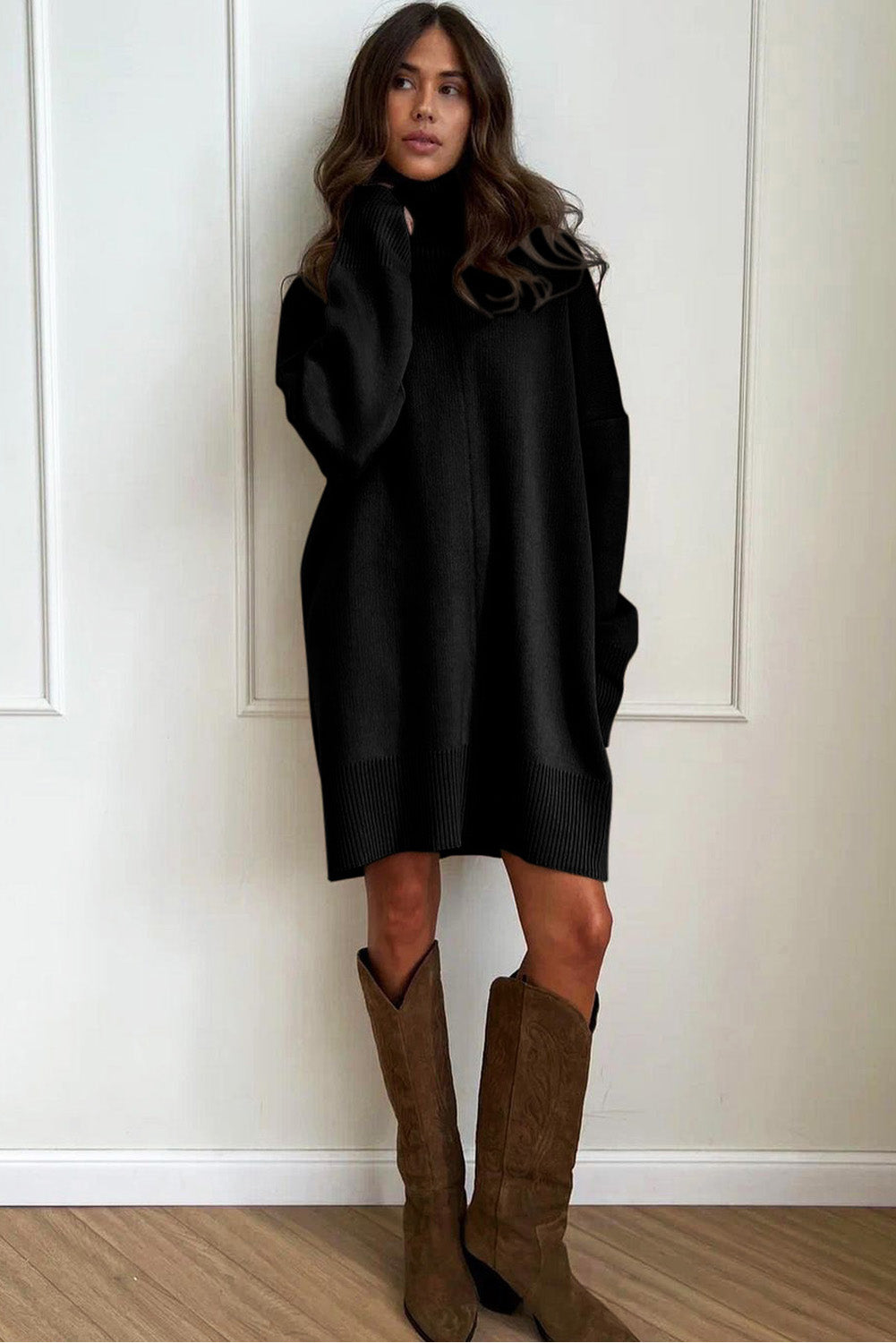 Woman wearing a black Oversized Turtleneck Jumper Dress and brown boots against a white wall.