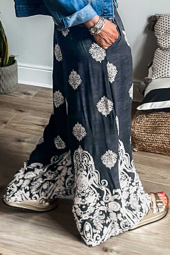 Women's black geometric print wide leg trousers in plus size with paisley border, styled casually with sandals and denim jacket indoors.