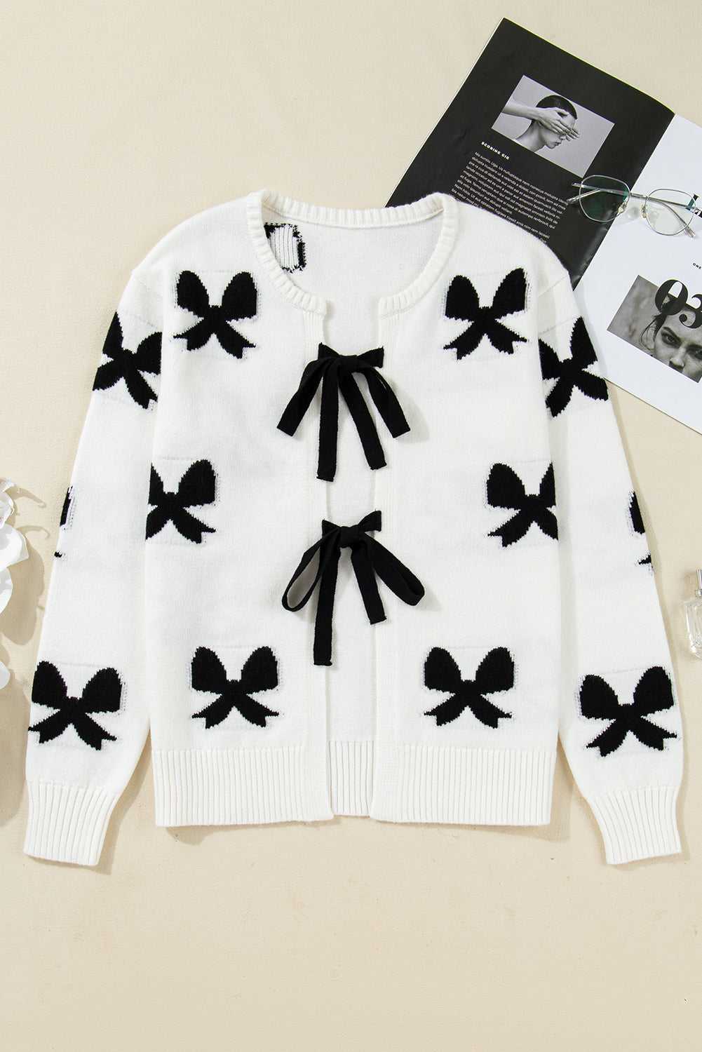 Flat lay of White Bow‑Lace Knit Cardigan with black bow patterns and ribbon ties, highlighting its soft knit texture and elegant design.