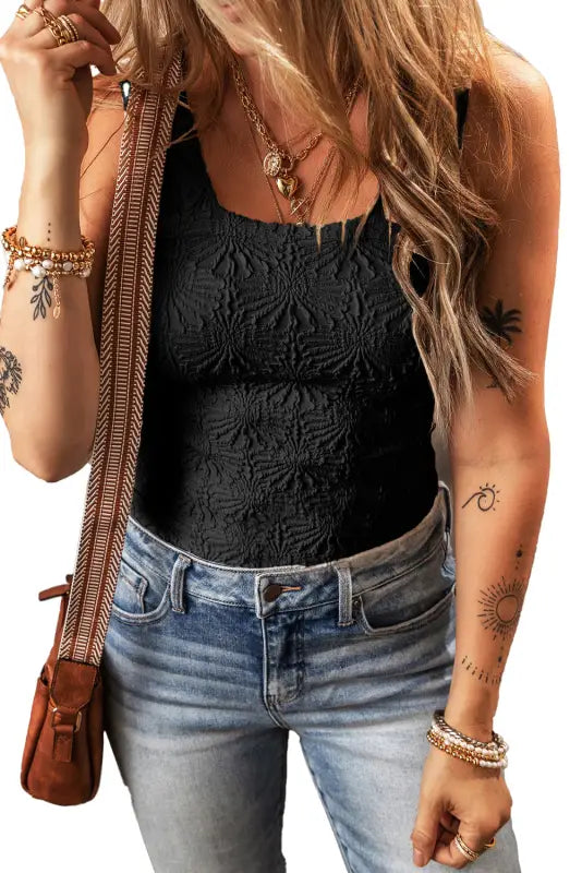 Woman in black floral jacquard tank top, light jeans, showcasing jewelry and tattoos