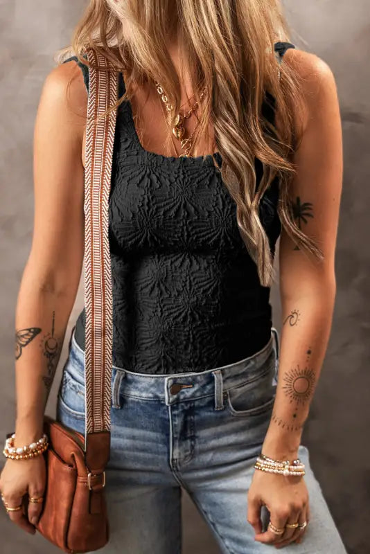 Woman wearing black floral jacquard tank top, blue jeans, jewelry, and brown shoulder bag