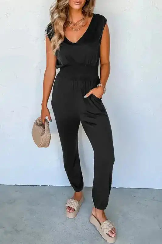 Black Shirred Jumpsuit V-Neck with high waist and sleeveless design, styled casually with beige woven sandals and clutch.