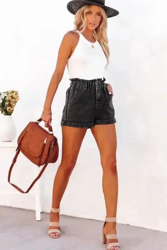 Full body view of model in black vintage washed denim shorts with frilled hem, styled with a white top, heels, and brown handbag.