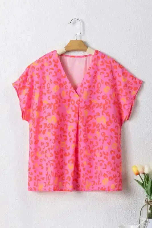 Flat lay of Wild in Pink Leopard Blouse on hanger. V-neck short sleeve top with bold pink, orange, and yellow leopard print for versatile styling.