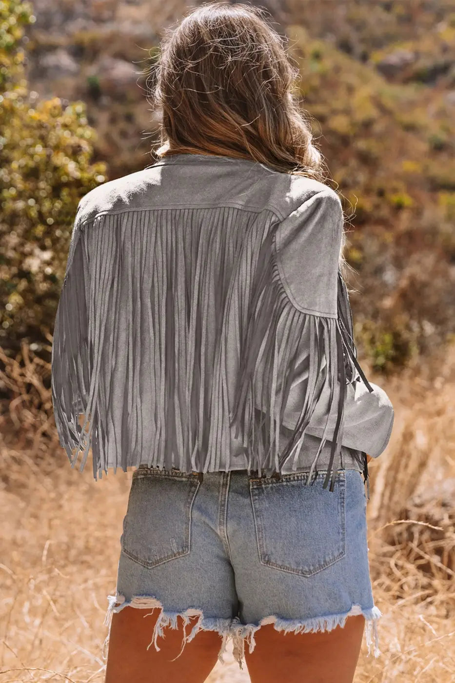 Back view of gray cropped fringe faux suede jacket paired with distressed denim shorts, showcasing full fringe movement.