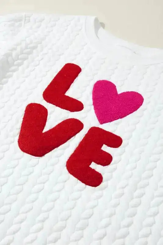 Detail of white cable knit sweatshirt with red 'LOVE' and pink heart chenille embroidery, perfect for seasonal gifting.