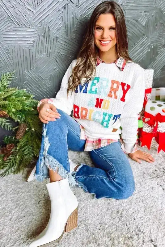 Model wearing white 'MERRY AND BRIGHT' chenille sweatshirt, paired with frayed jeans and white boots for holiday outfit inspiration.