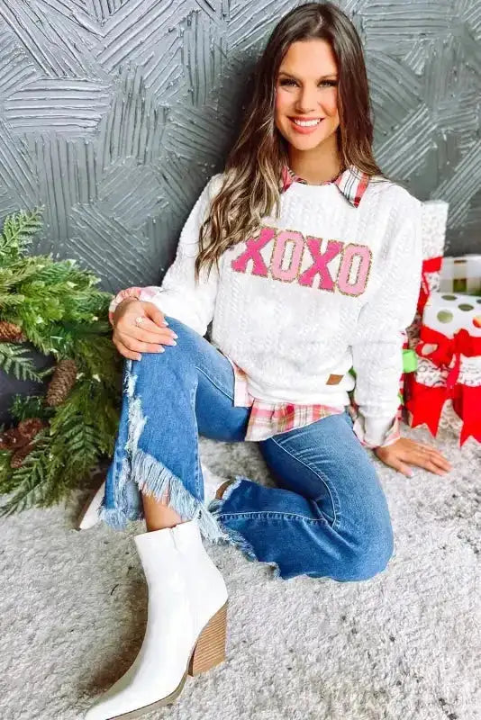 Model in white 'XOXO' chenille sweatshirt with pink and gold embroidery, paired with jeans and boots for cozy seasonal style.