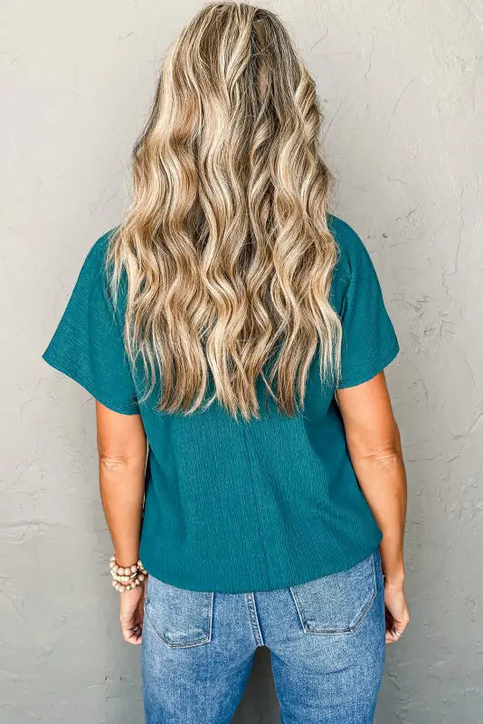 Long wavy blonde hair, teal shirt, blue jeans, wearing black crinkled v neck wide sleeve t-shirt