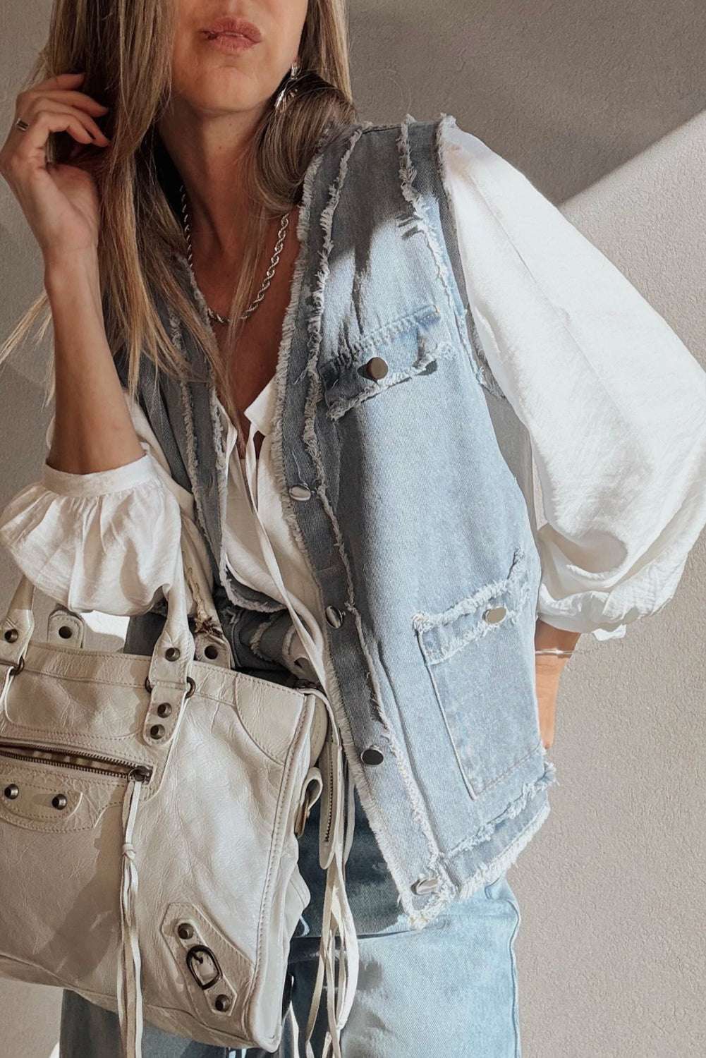 Front view of Beau Blue Denim Waistcoat with Frayed Edges worn with a white blouse and accessorized with a white handbag for a modern casual look.