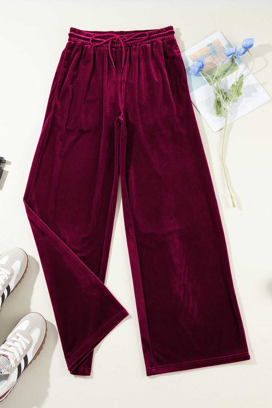 Flat lay of burgundy velvet yoga pants with wide-leg cut and drawstring waist, styled with sneakers and floral props for winter fashion.