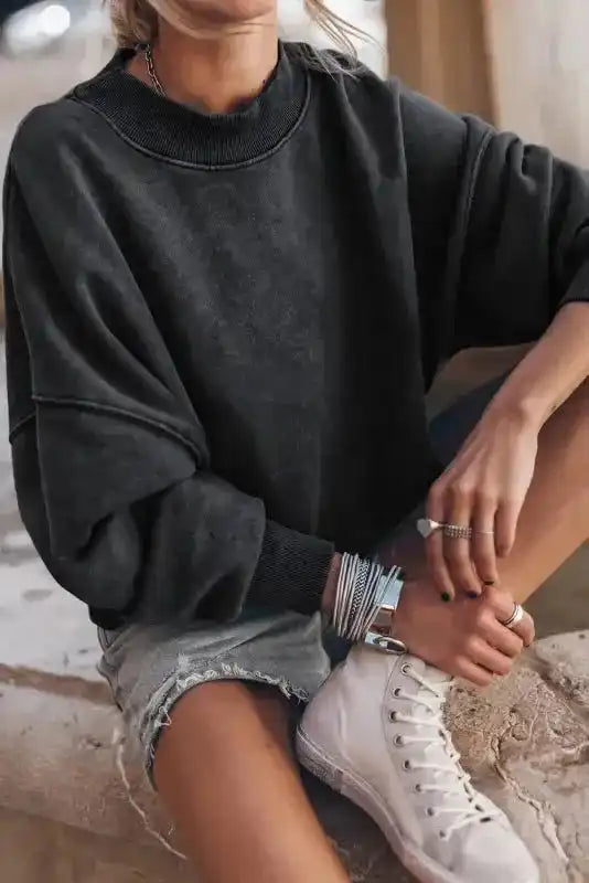 Seated model in black Cozy Drop Shoulder Crew Neck Sweatshirt with rolled sleeves and sneakers, styled for casual winter fashion.