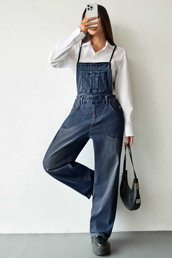 Dark blue baggy denim overalls for women with adjustable straps and front pockets. Casual denim jumpsuit styled with a white shirt and black handbag.
