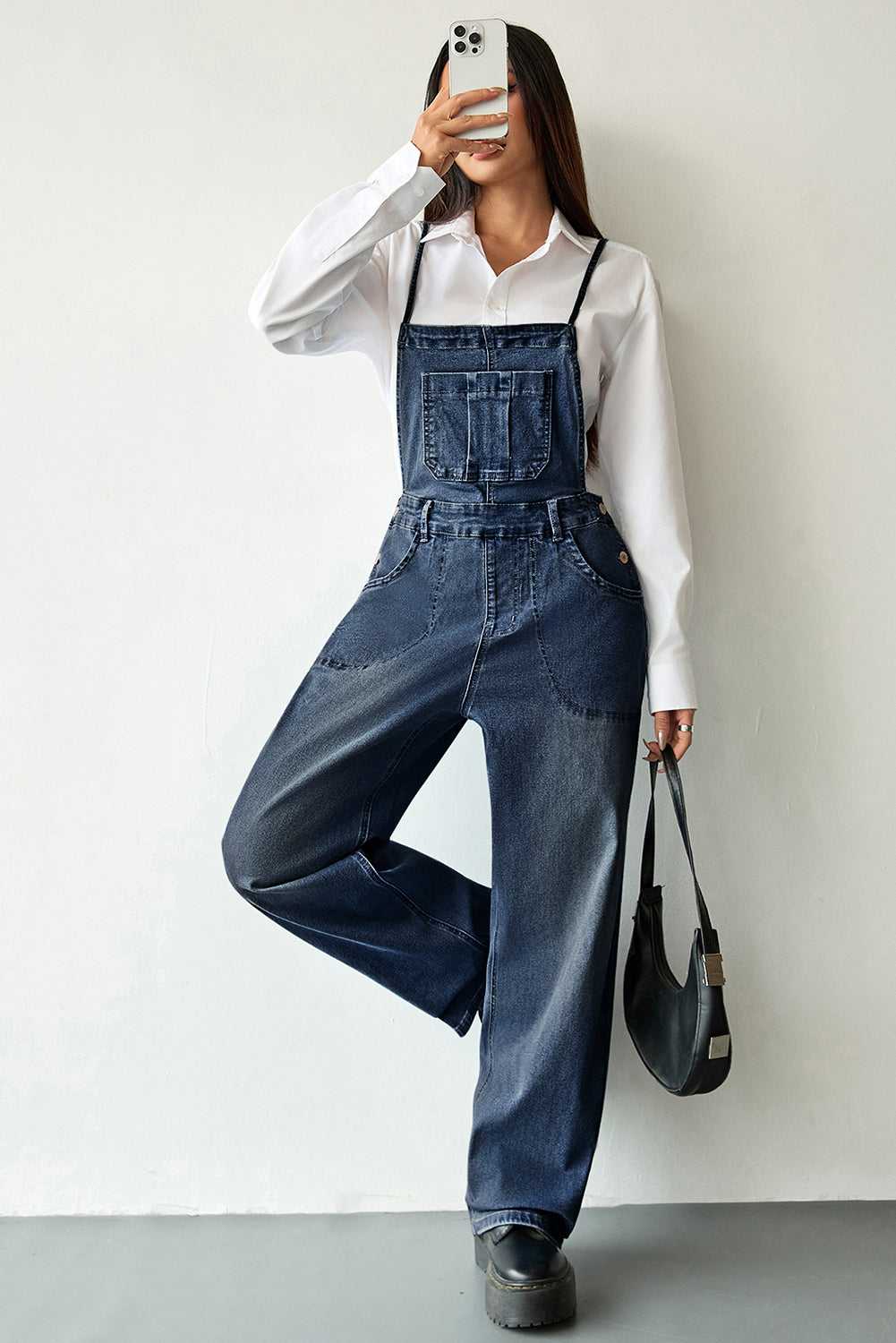 Dark blue baggy denim overalls for women with adjustable straps and front pockets. Casual denim jumpsuit styled with a white shirt and black handbag.