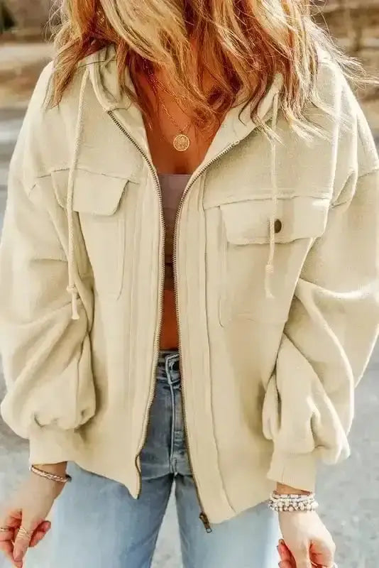 Front close-up of beige zip up hooded jacket women with button flap pockets and ribbed cuffs. Soft fleece fabric for warmth.