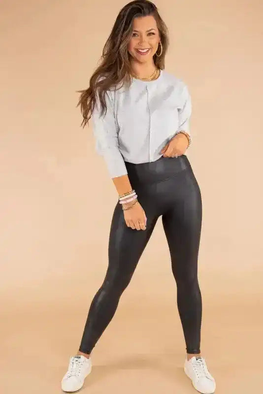 Full-body front view of model wearing women’s black faux leather skinny leggings with high-rise waist, styled with casual top and sneakers.