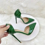 Pair of croc-effect green stilettos held above white platform, showcasing elegant heel height and fashionable cut-out sides.