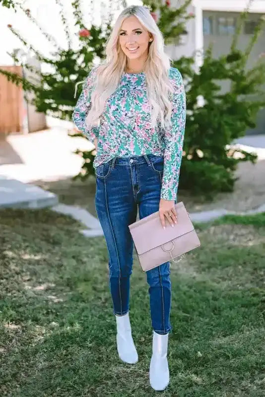 Full body view of woman in green floral long sleeve top and skinny jeans, accessorized with white boots and a pink clutch.