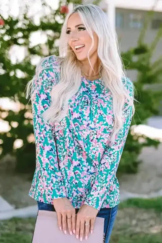 Smiling model wearing green floral long sleeve top with pink accents, paired with jeans and a blush clutch.
