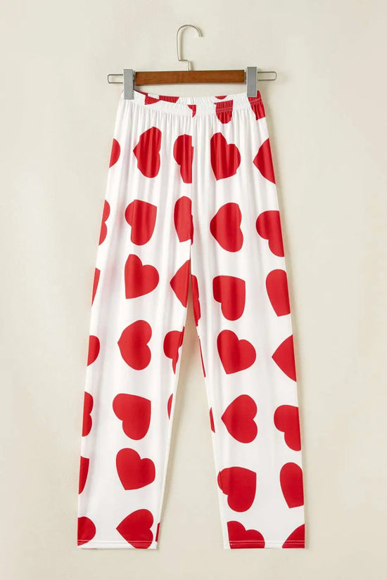 White pants with red heart patterns hanging on a wooden hanger against a beige wall.