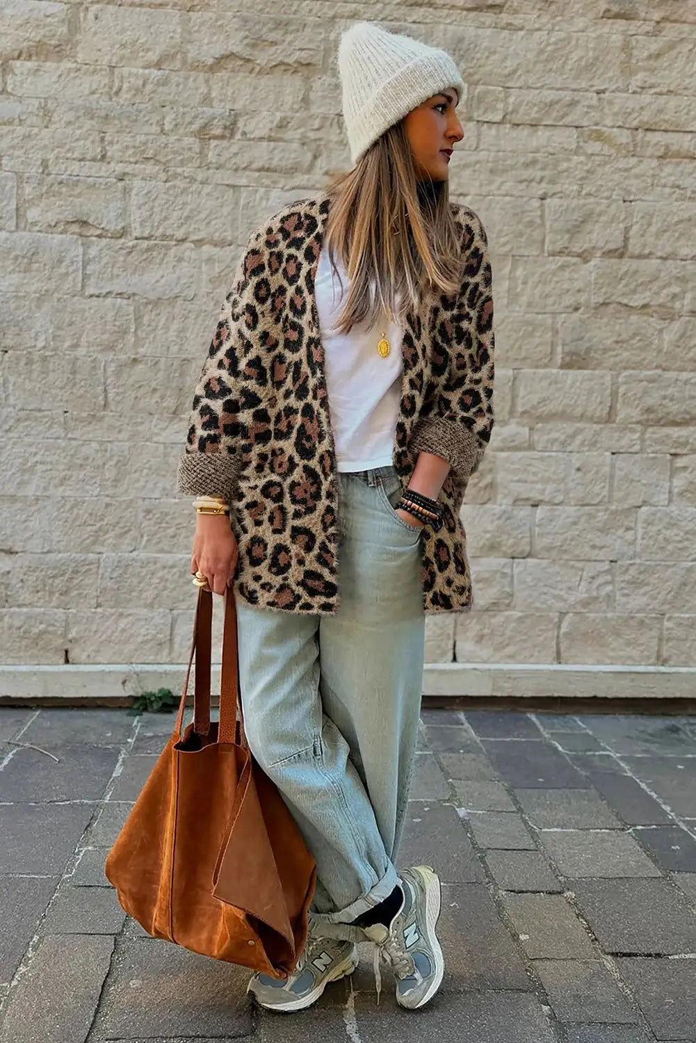 Person wearing a leopard print coat, white beanie, light blue jeans, and holding a brown leather bag against a stone wall.