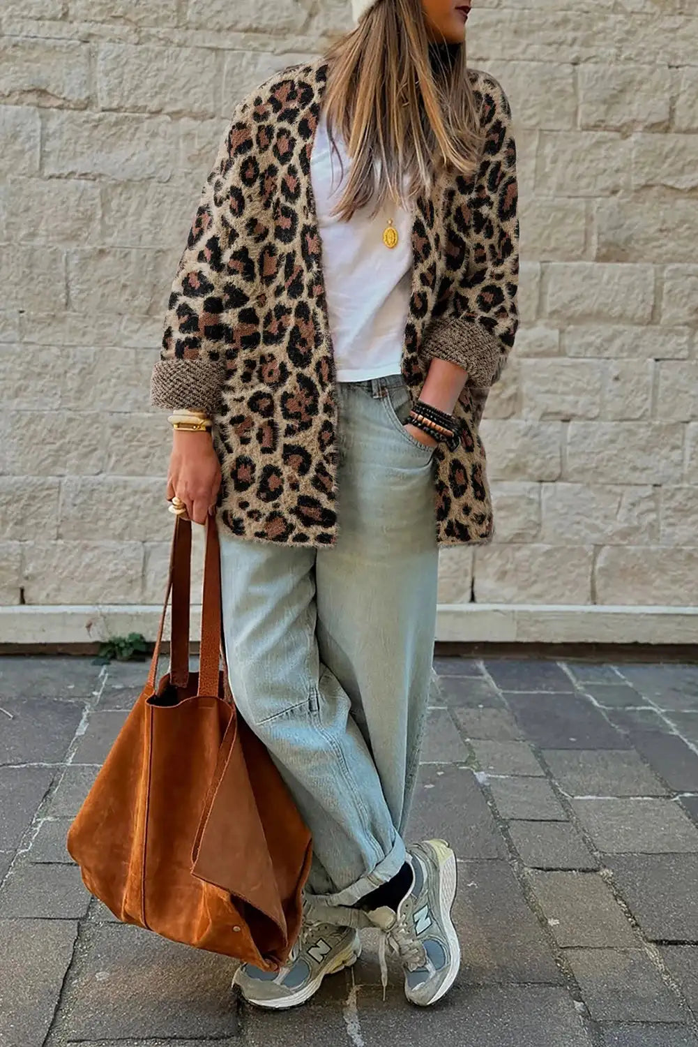 Person wearing a leopard print coat, light blue jeans, and holding a brown bag against a stone wall.