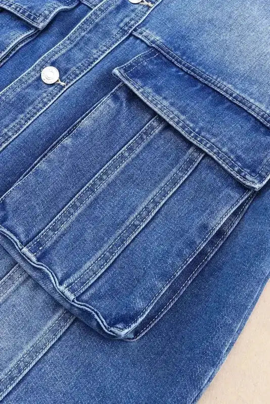 Close-up of oversized chest pocket on women's blue denim jacket showing structured pleats and solid color fabric.