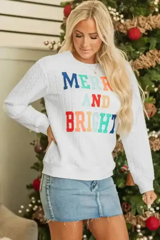 Model wearing white Merry And Bright Cable Knit Sweatshirt with colorful text, styled for holiday fashion.