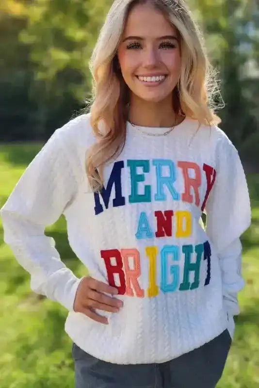 Smiling woman wearing white Merry And Bright Cable Knit Sweatshirt outdoors, showcasing cheerful holiday apparel.
