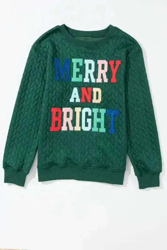 Flat lay of Merry And Bright Cable Knit Sweatshirt | FashionFitz showing colorful embroidered text on green quilted fabric.