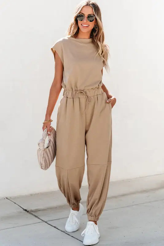 Beige open back drawstring jogger jumpsuit with sunglasses, handbag, and white sneakers