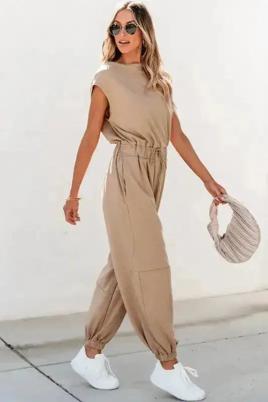 Side walking pose in beige jogger jumpsuit with cap sleeves and tapered legs, combining comfort and chic style.