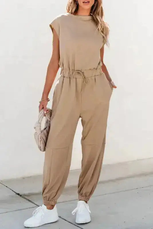 Open Back Jogger Jumpsuit – Beige with cap sleeves and drawstring waist, styled with white sneakers for casual women’s wear.